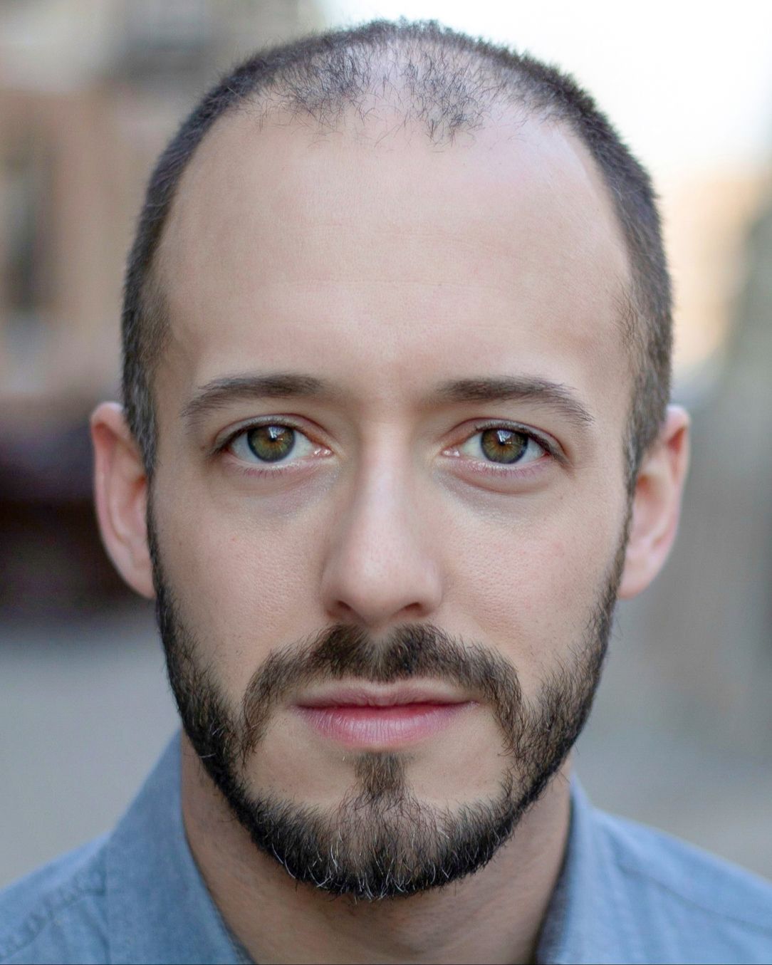 Professional headshot of a person in a blue shirt against a blurred urban setting.