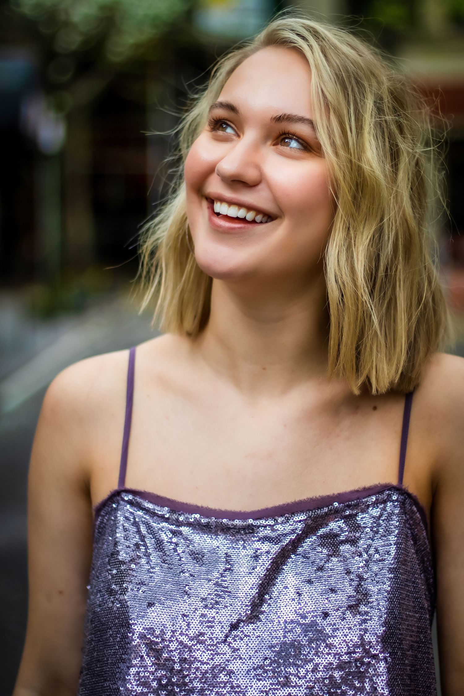Candid portrait of someone in a sparkly purple top with short blonde hair laughing outdoors.