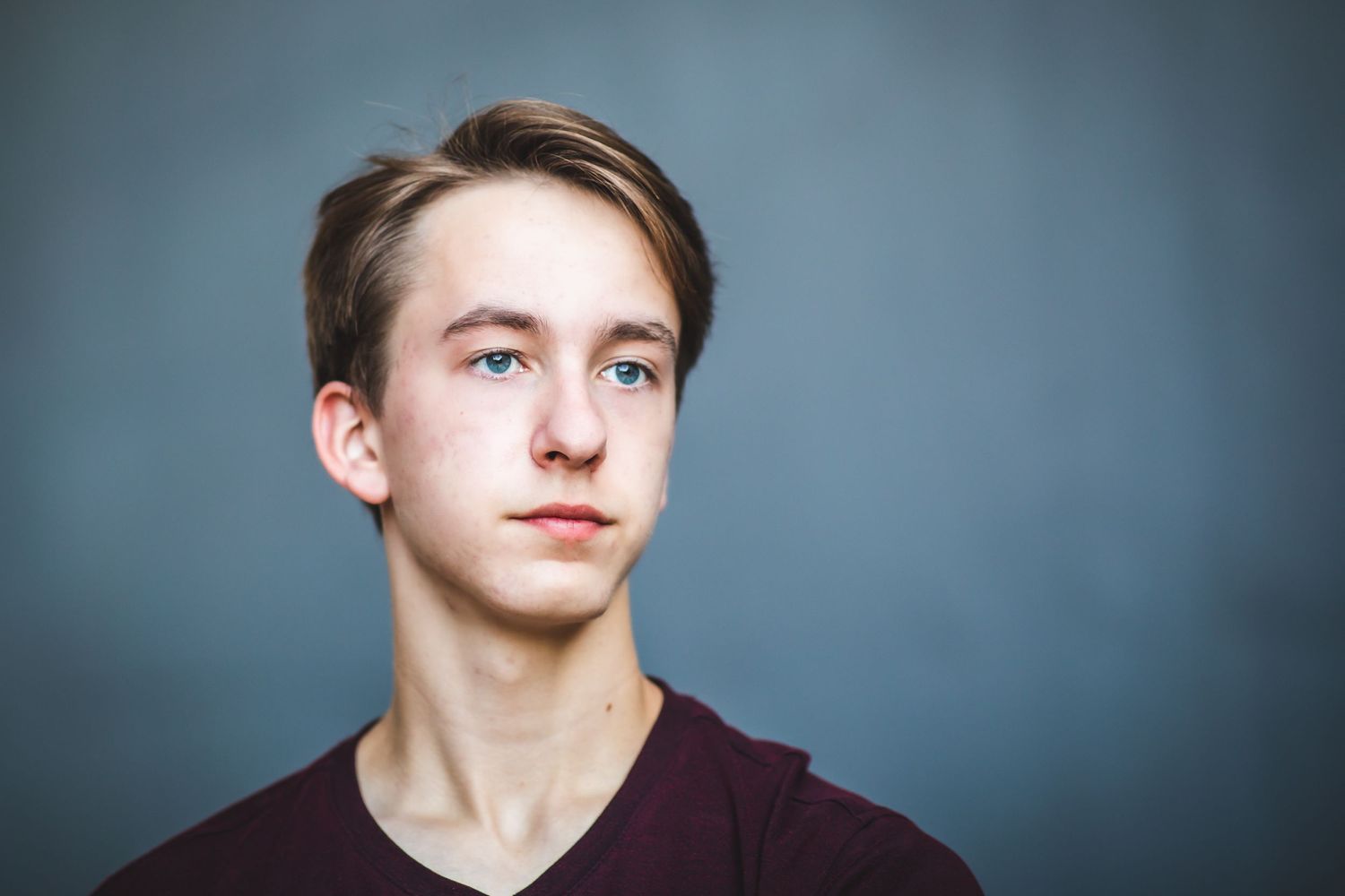 Young dancer in burgundy shirt poses against grey background in studio portrait.