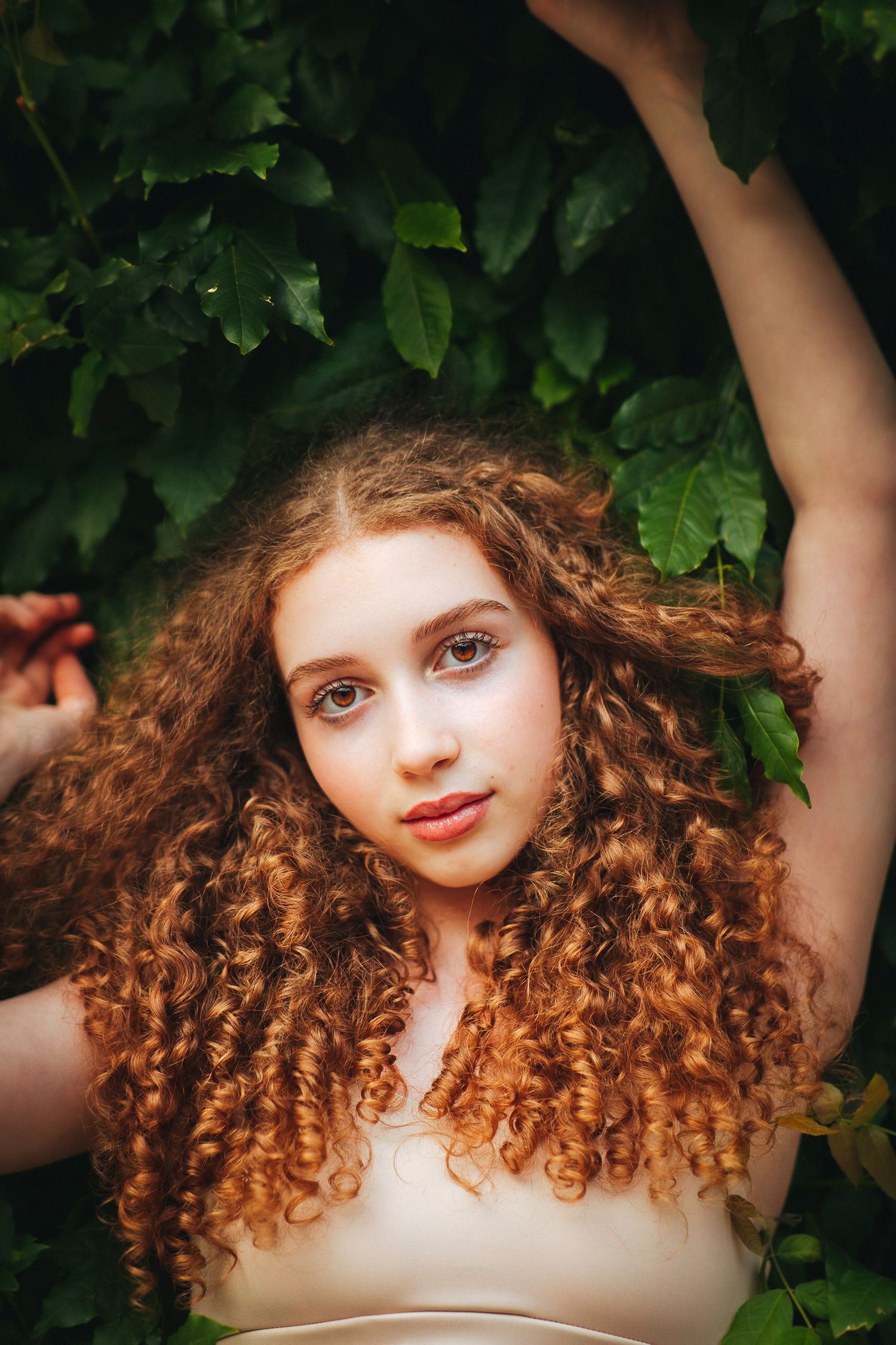 Portrait of a person with flowing curly auburn hair posing against green foliage in natural lighting.