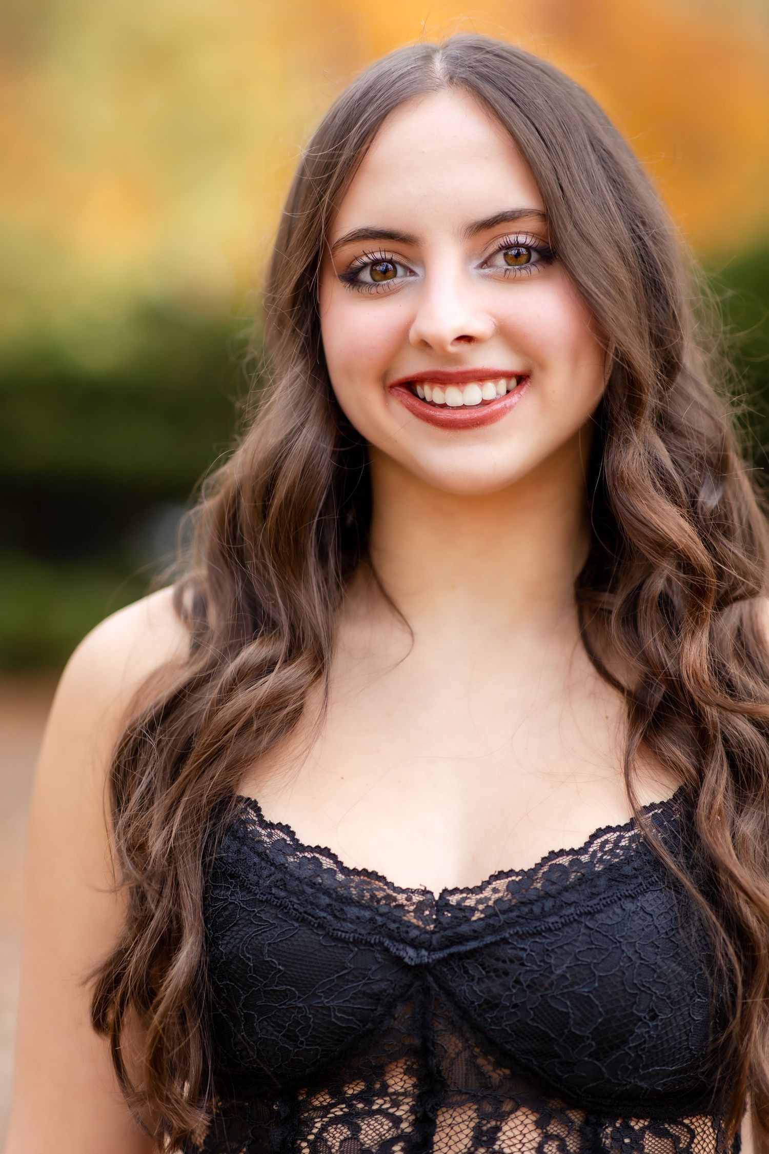 Portrait of a smiling young woman with long wavy dark hair in black lace top against autumn backdrop.