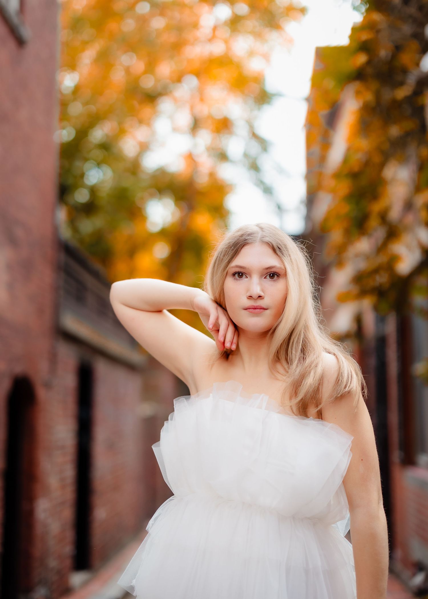 Portrait in white dress against brick wall with autumn foliage.