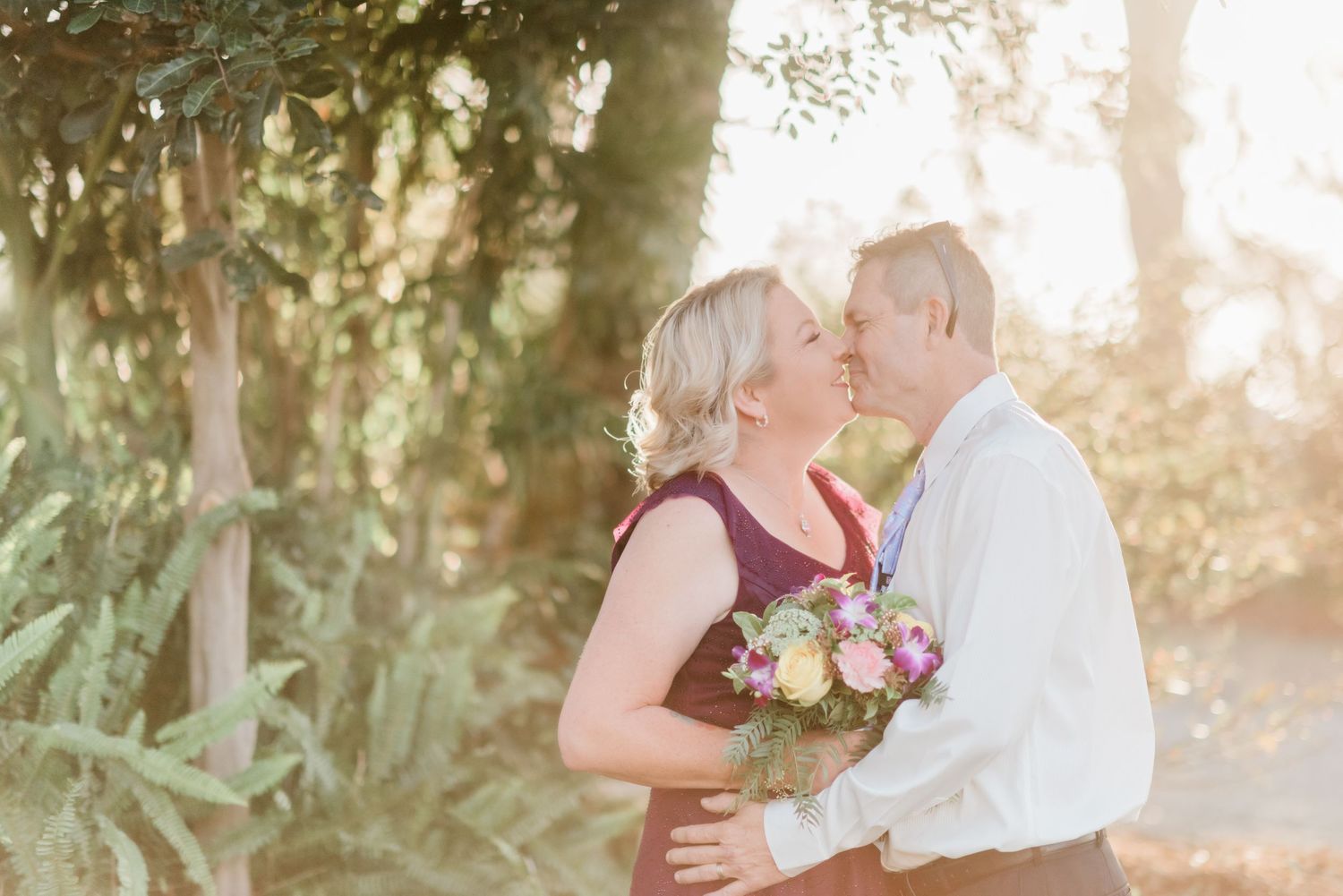 Romantic outdoor couple portrait with sunlit background and floral bouquet.