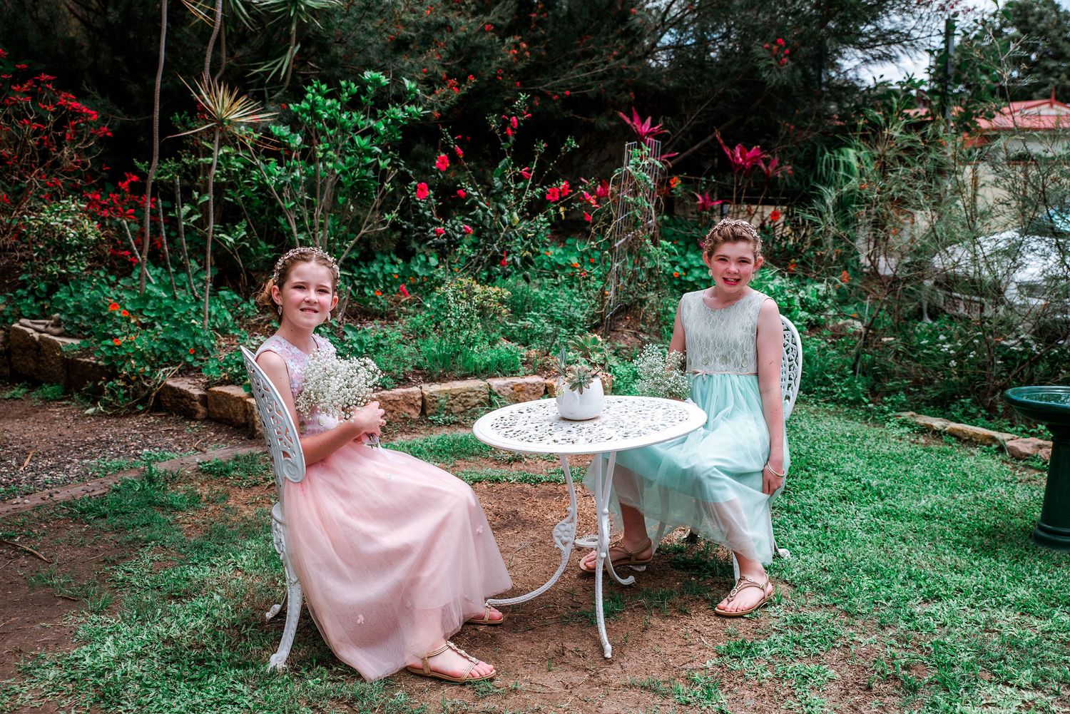 Two people in vintage-style dresses enjoy tea at an outdoor garden table among blooming flowers.