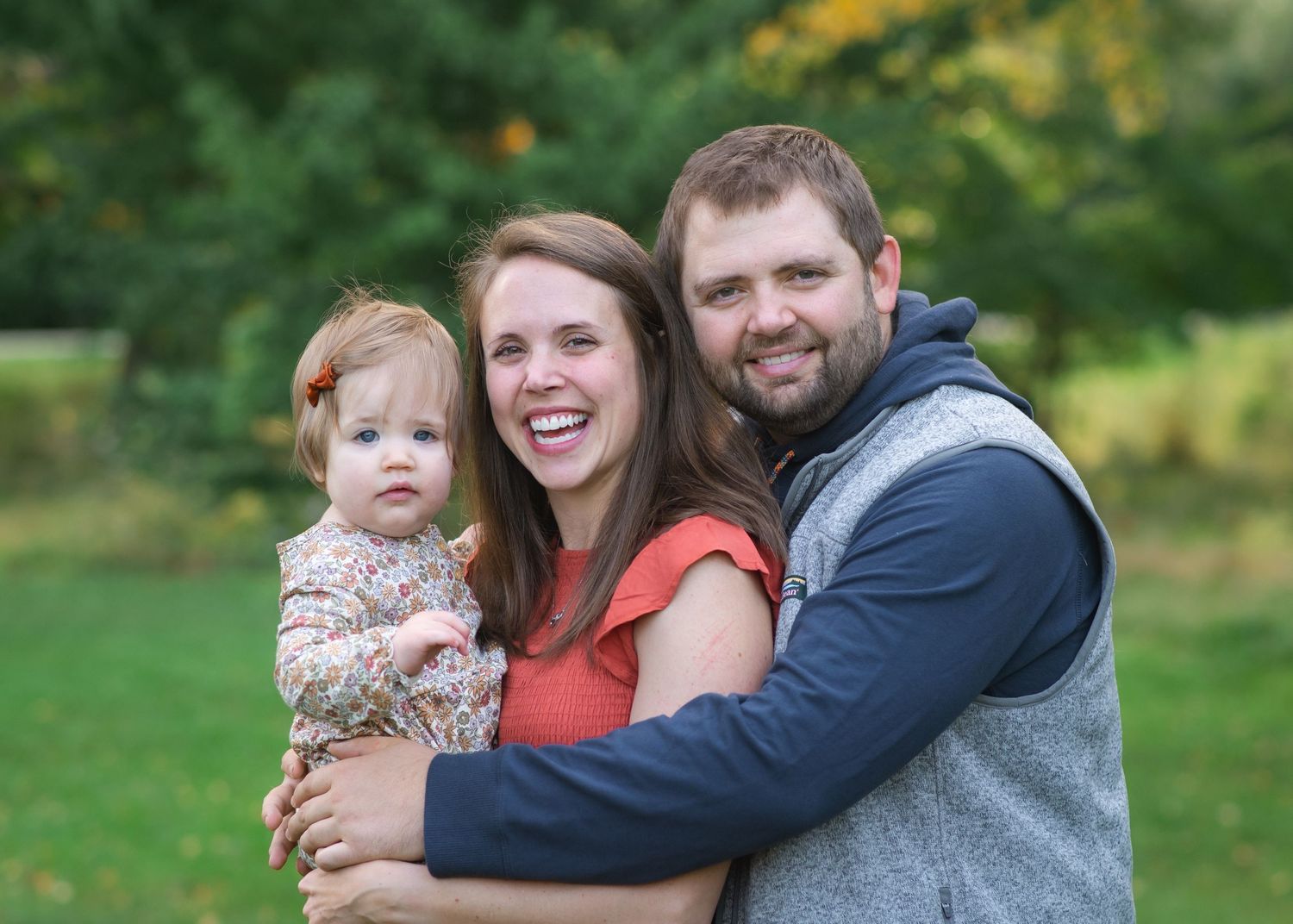 An outdoor family portrait shows affectionate embrace against a green natural background.