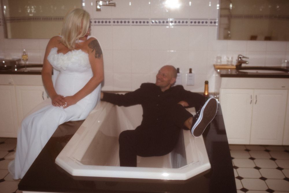 Couple poses playfully in white bathtub during wedding photoshoot.
