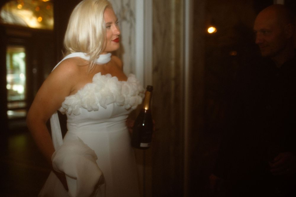 Woman in white wedding dress holding a bottle in dimly lit setting.
