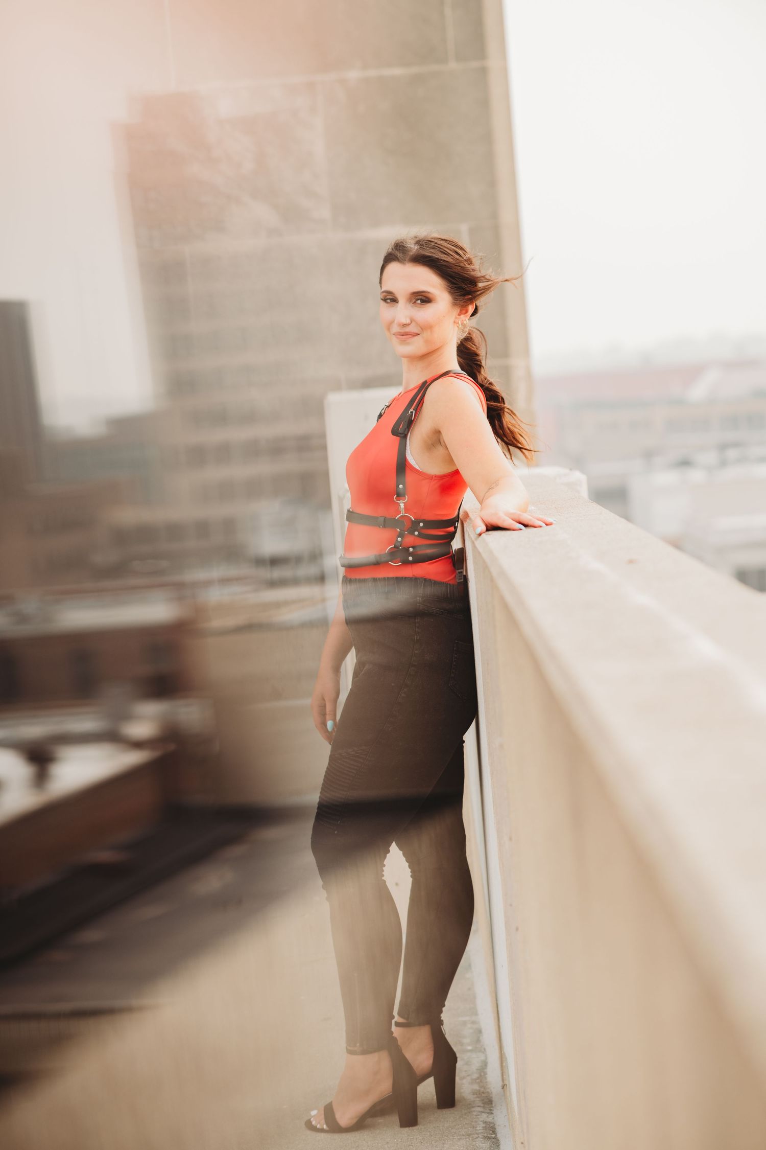Woman in coral top and dark pants poses on urban rooftop during sunset.