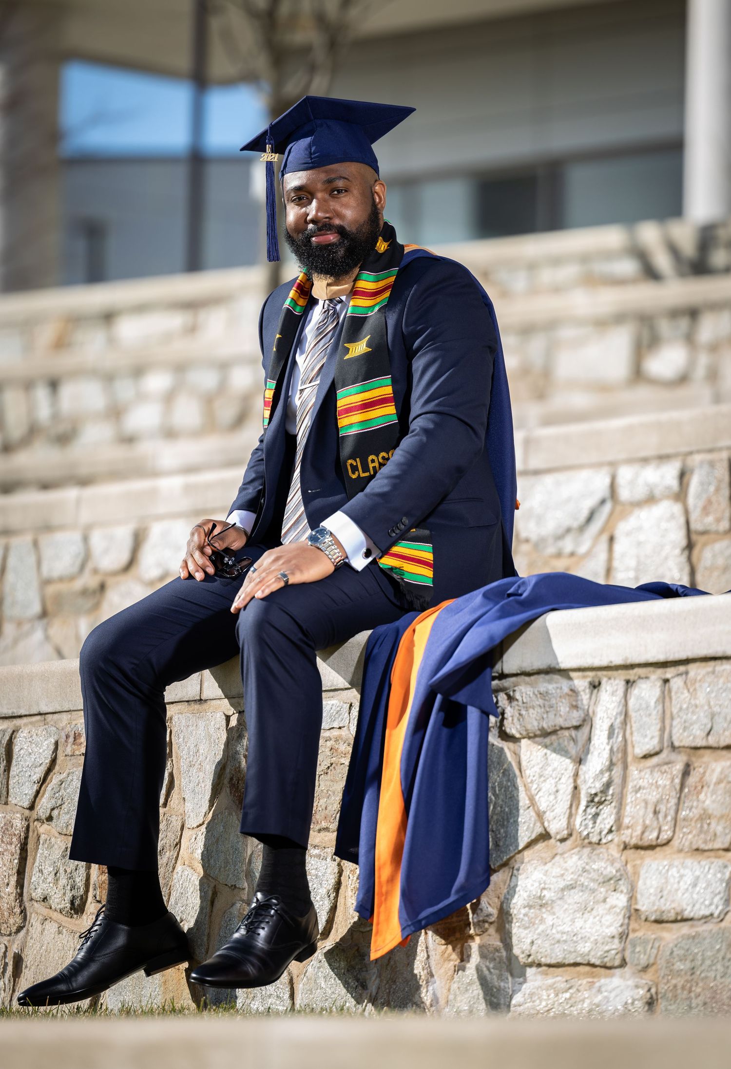 Graduate in navy cap and gown with kente stole sits casually on stone steps.