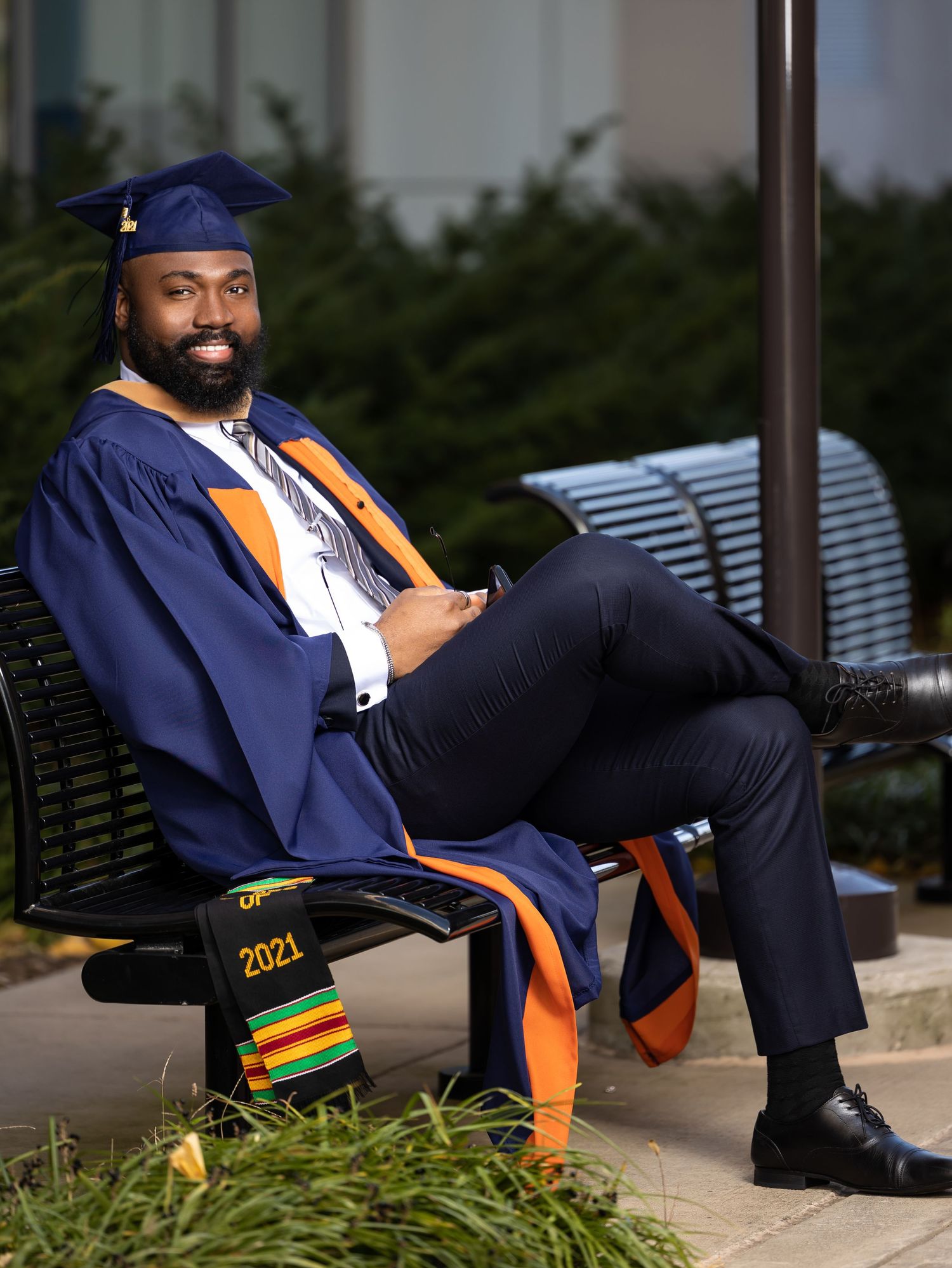 Graduate in academic regalia displays Class of 2021 kente stole while seated outdoors.