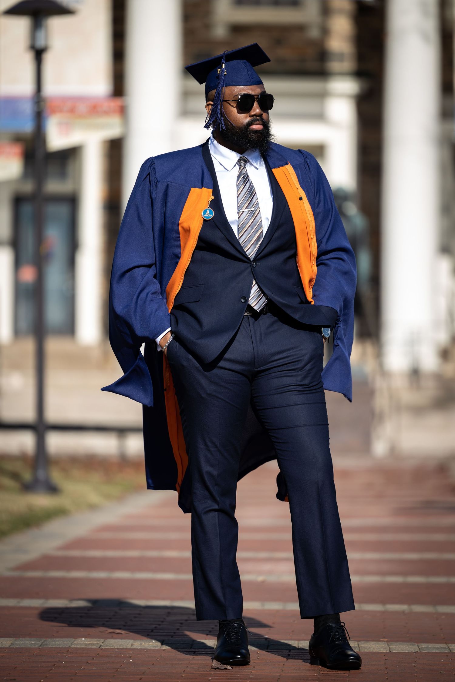 Graduate in sunglasses poses stylishly in navy graduation gown with orange accents.