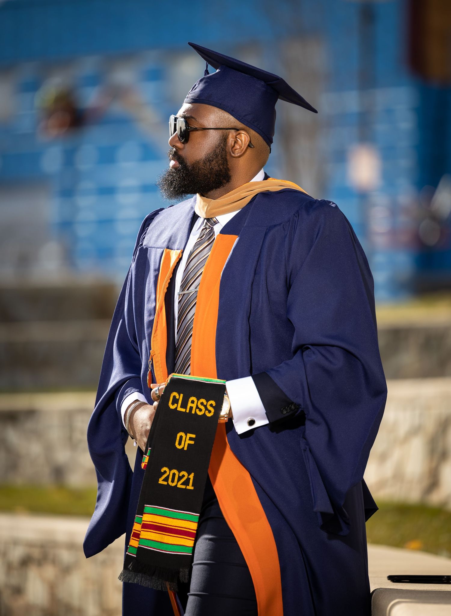 Graduate in navy blue regalia displays Class of 2021 sash with kente cloth accents.
