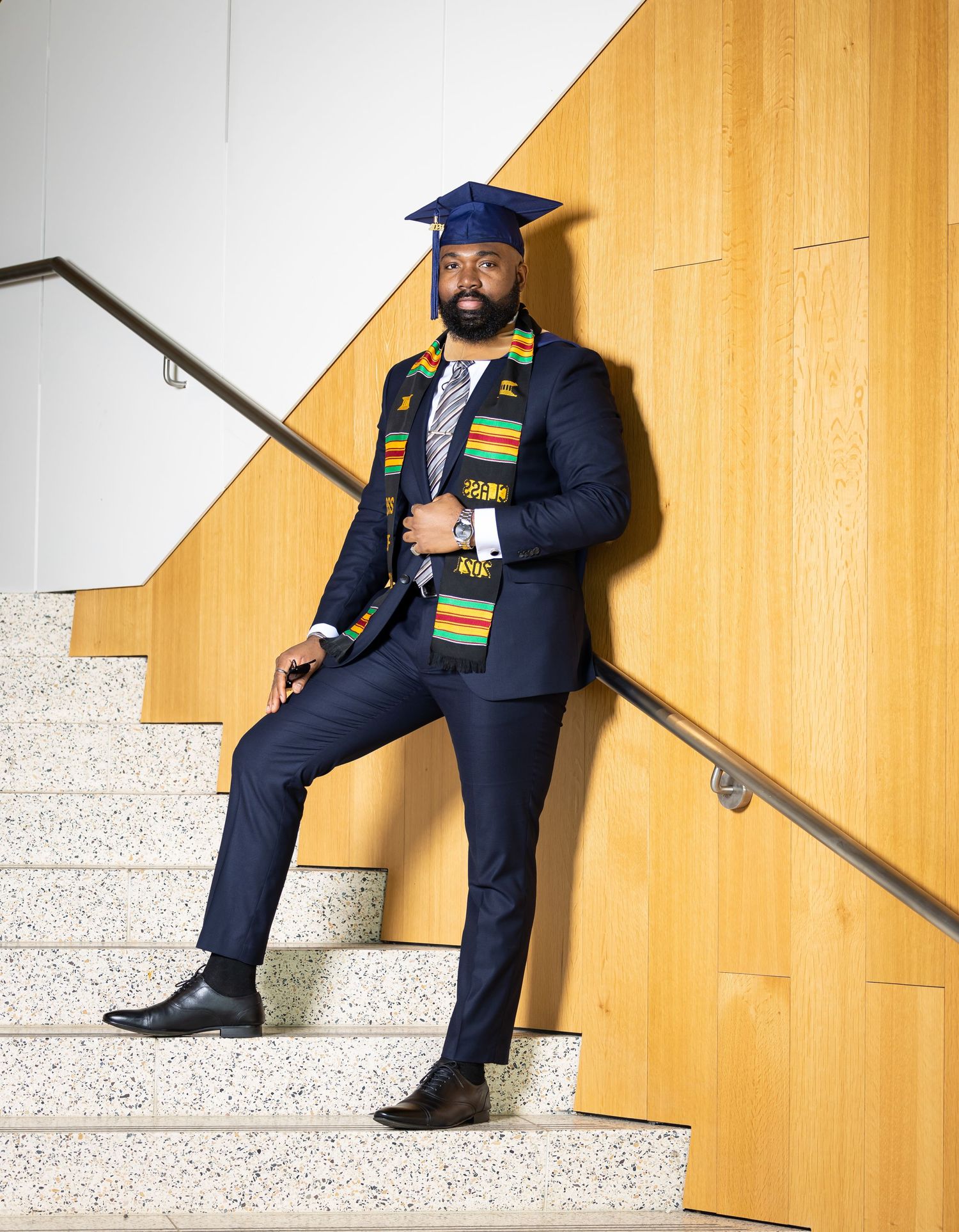 Graduate poses on stairs in academic regalia against golden wall with kente stole.