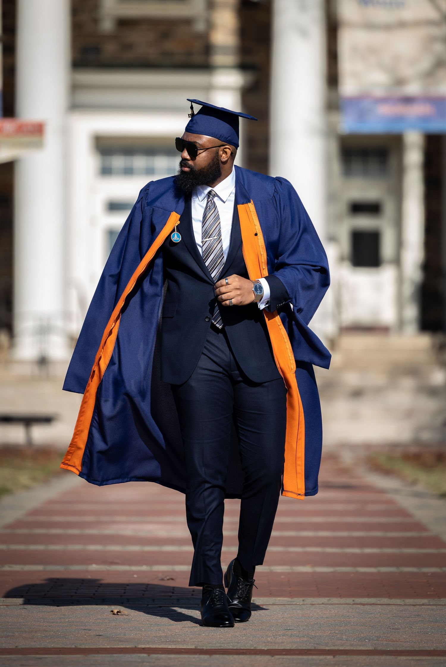 Graduate walks confidently in navy blue academic regalia with orange trim and kente stole.