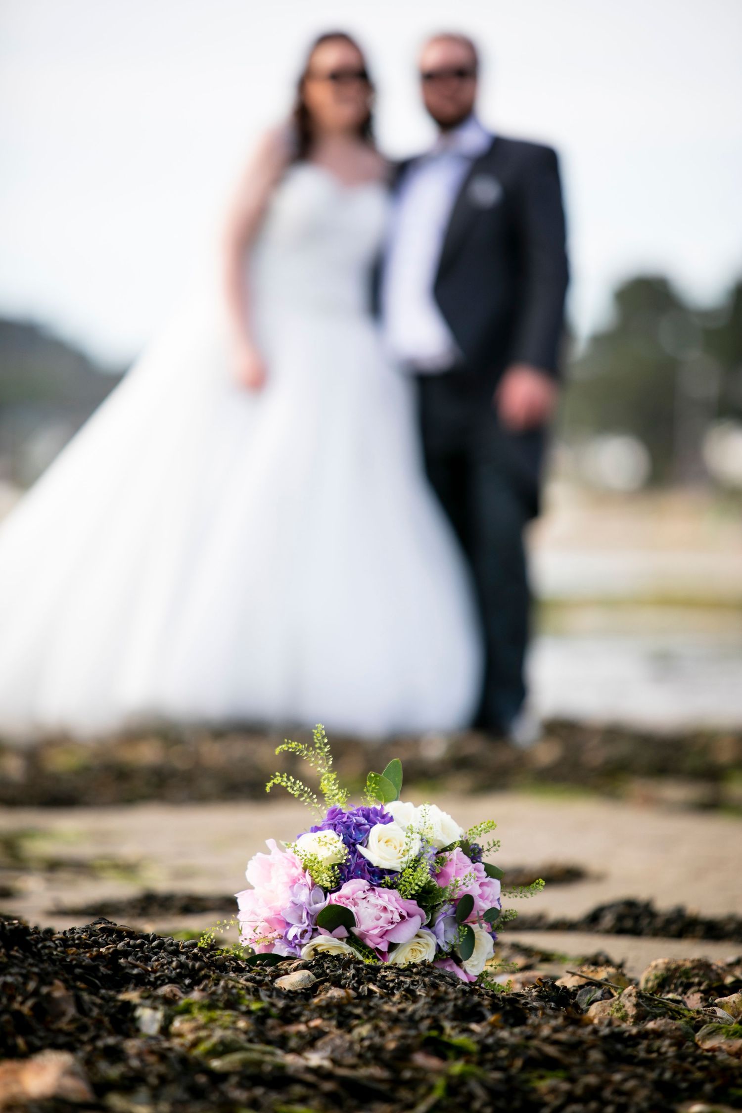 Wedding couple silhouettes with pink and purple floral bouquet in foreground on rocky beach.