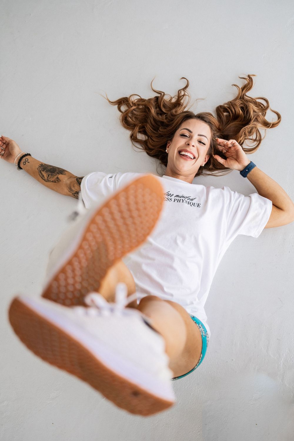 Person lying on white floor wearing white t-shirt and sneakers with hair spread out and laughing.