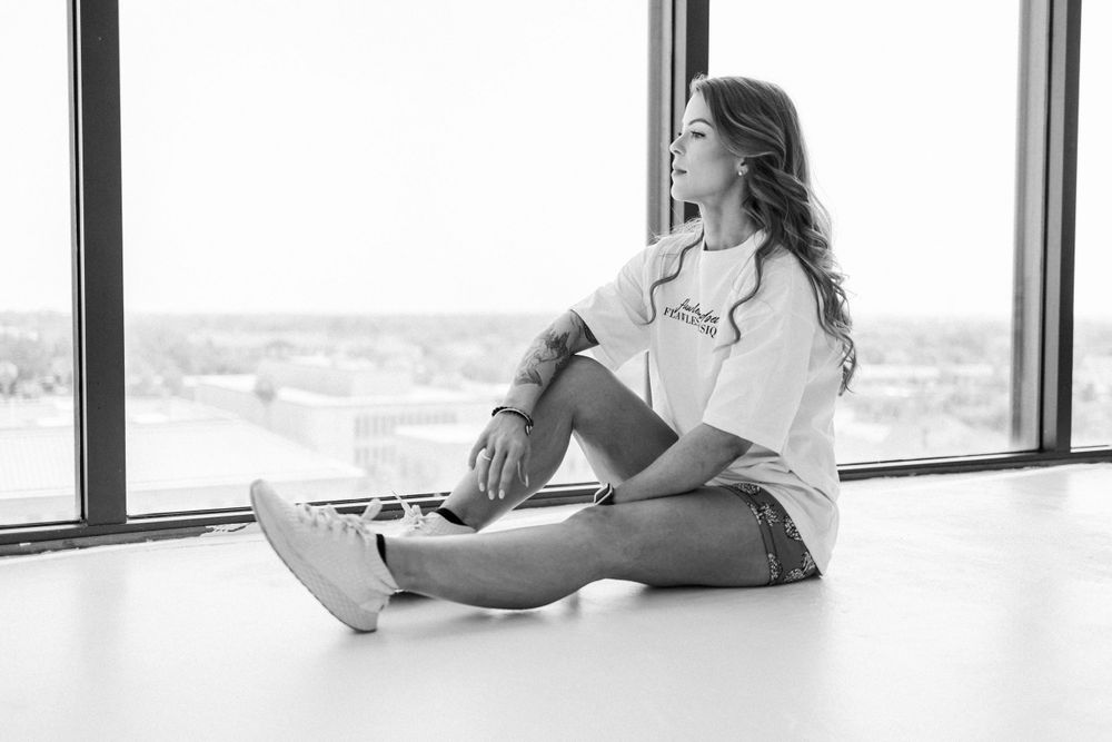 Monochrome photo of person sitting by window wearing casual attire and sneakers.