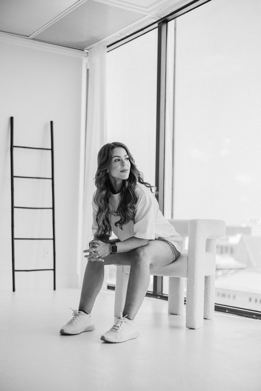 Model in white top and shorts sits thoughtfully by window in minimalist white room with decorative ladder.