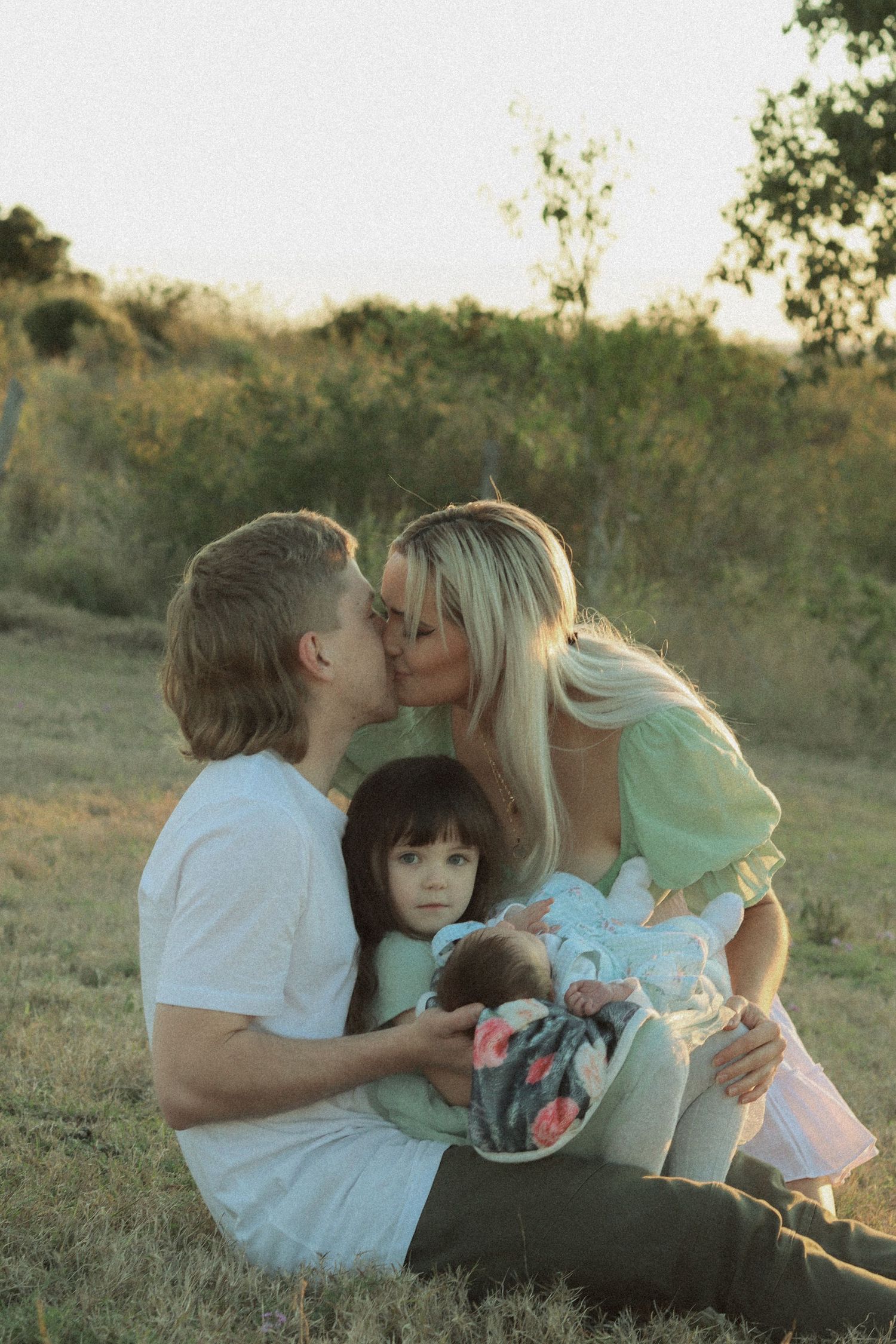 A tender family moment captured at sunset in a grassy field with soft golden lighting.
