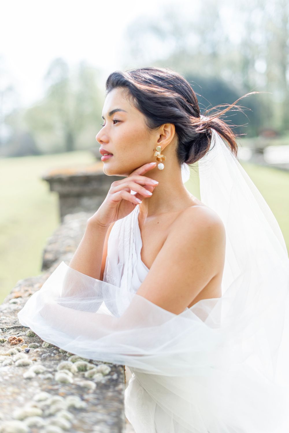 A bride in an elegant white off-shoulder gown with flowing sleeves poses outdoors wearing pearl drop earrings and a veil.
