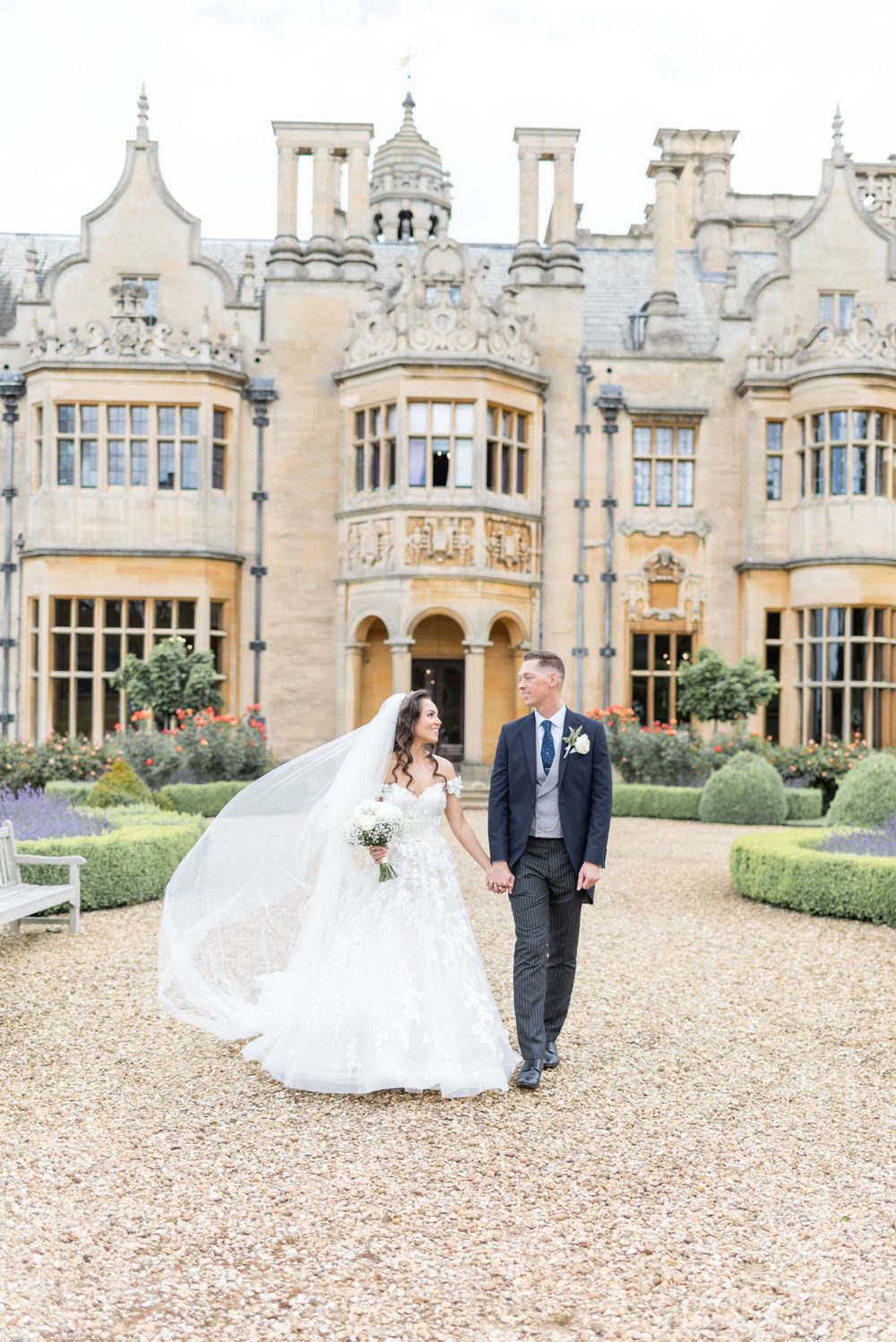 Elegant wedding couple standing outside historic Harlaxton Manor with ornate Gothic architecture and manicured gardens.