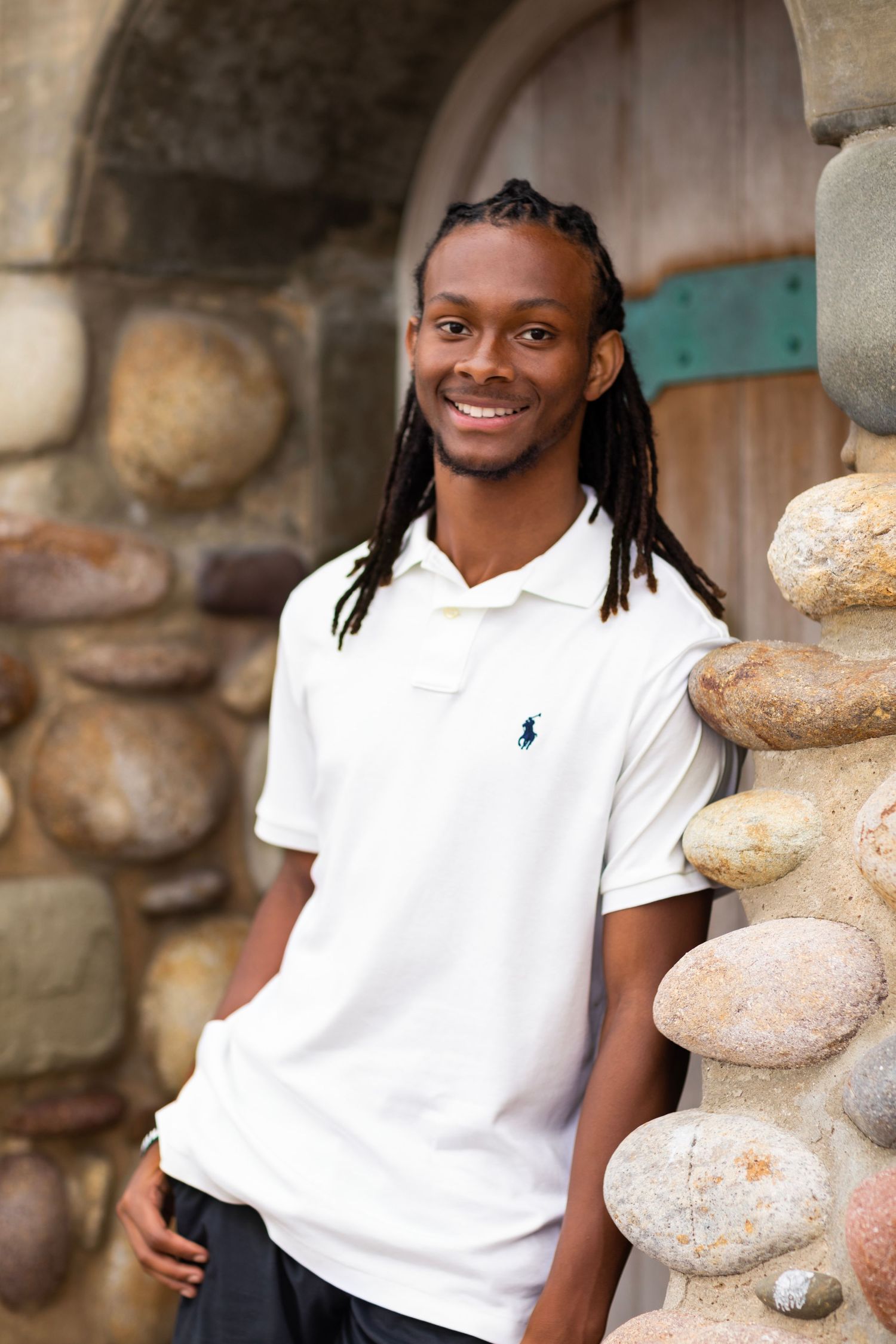 A high school senior in white polo shirt posing against stone wall with warm genuine expression.