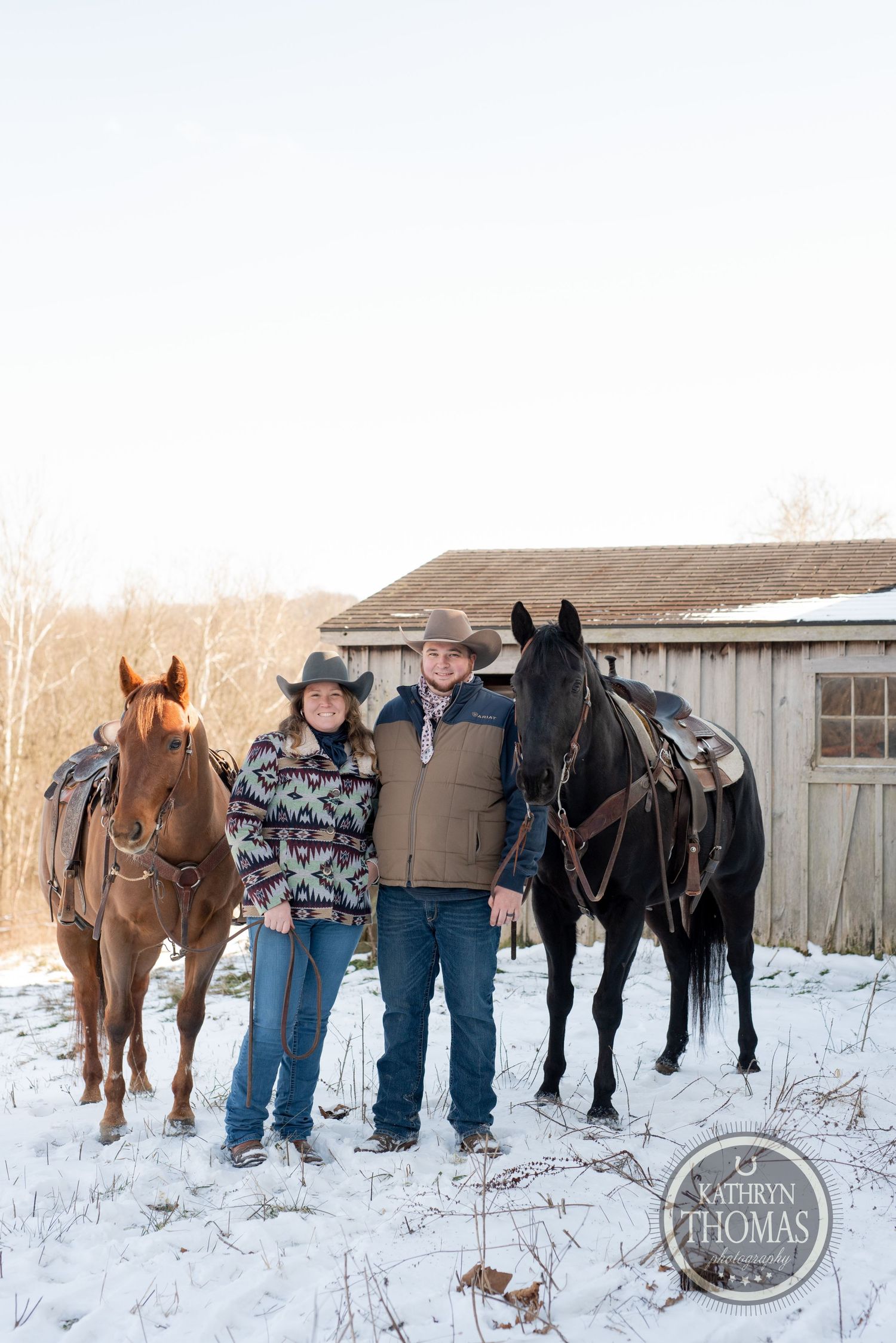 Two horses and their riders stand together in the snow by a weathered barn building.