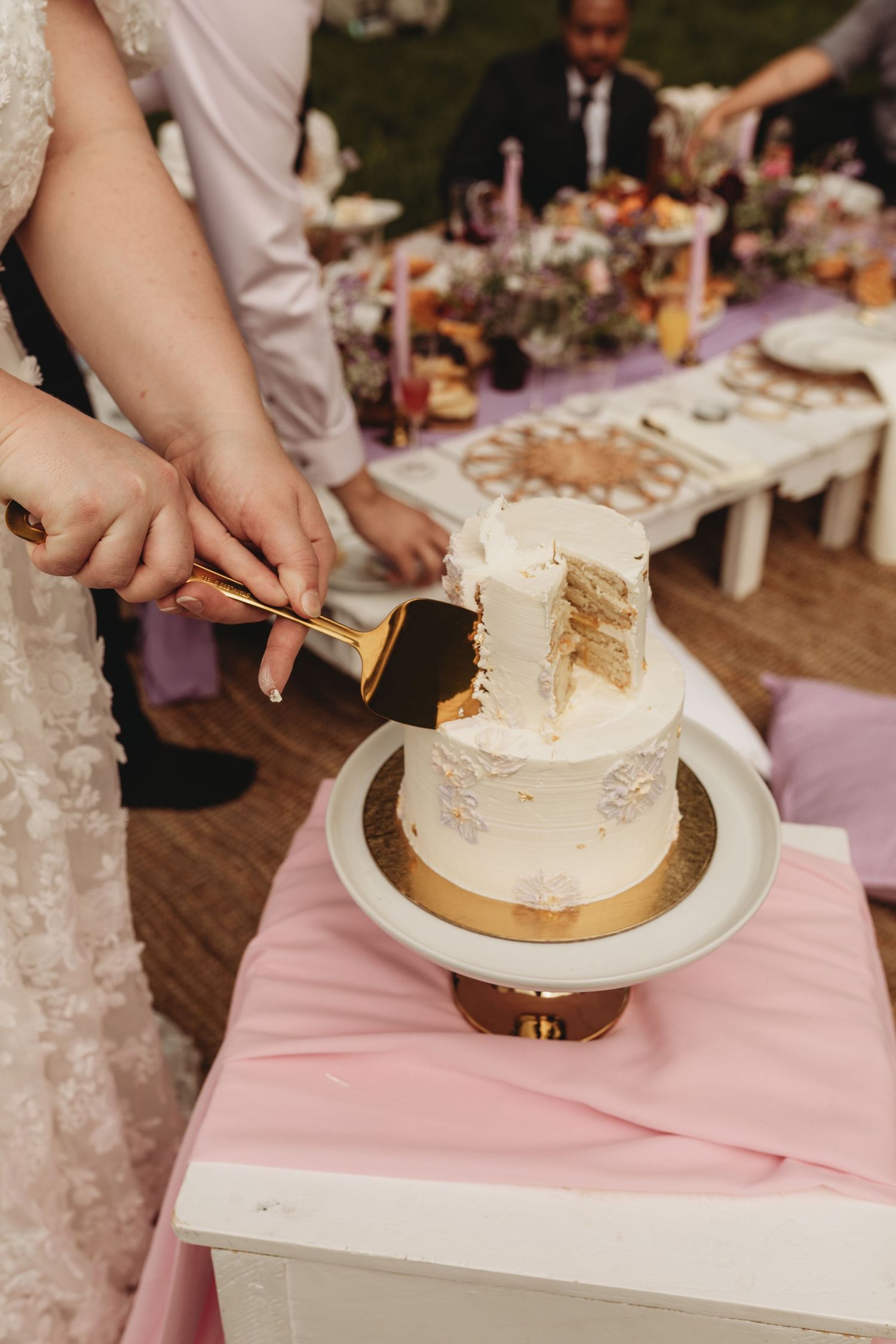 Close up of hands cutting into a white tiered wedding cake on a pink table setting.