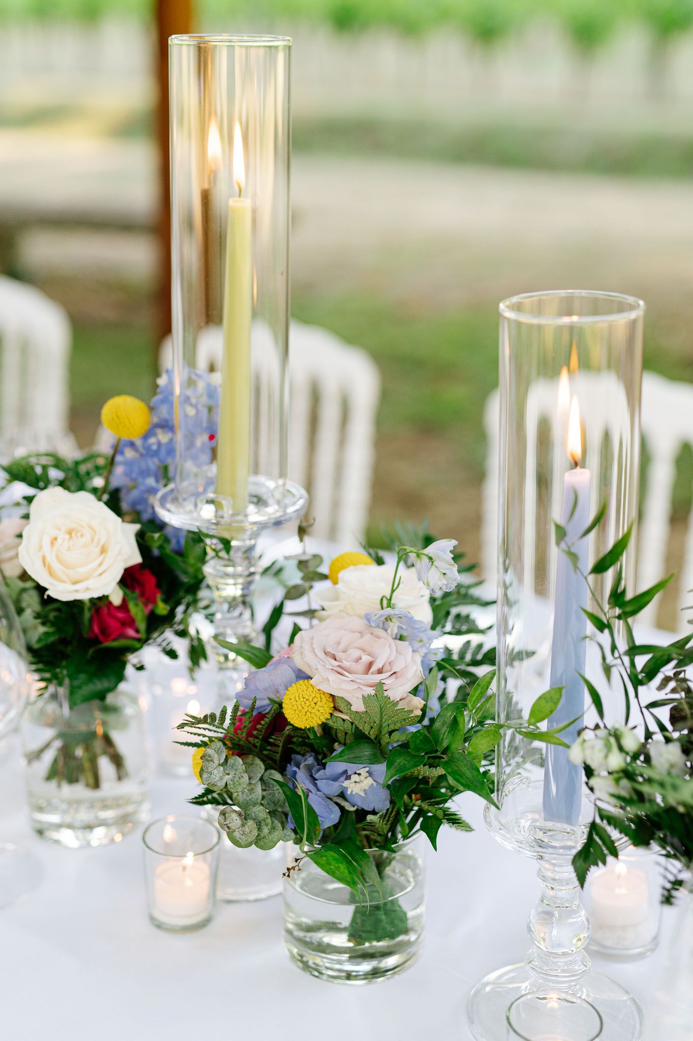 Wedding centerpiece with tall glass candle holders, roses and delicate blue flowers on white tablecloth.