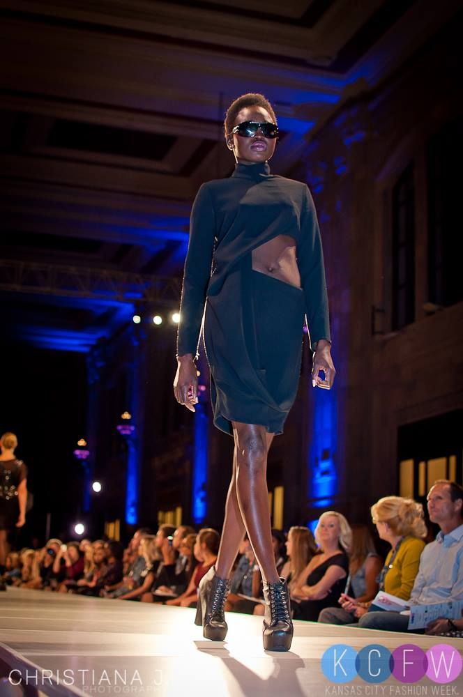 Fashion model walks runway in blue dress and sunglasses with dramatic lighting.