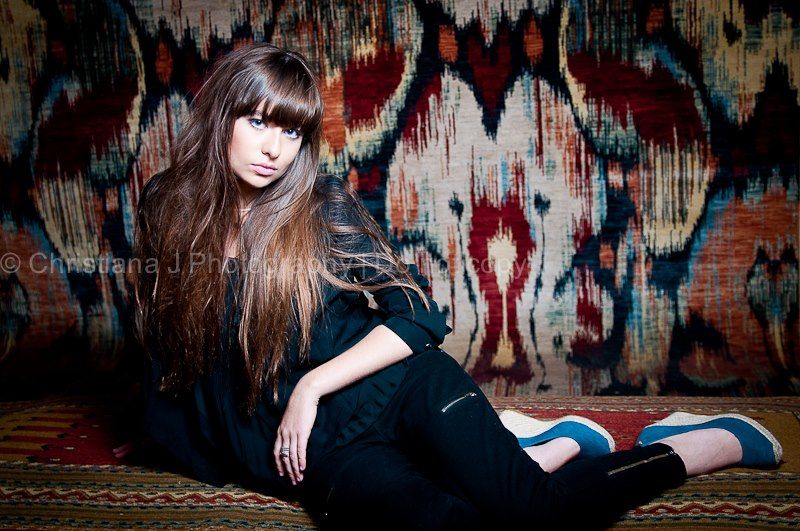 Fashion model lounging on patterned carpet wearing black outfit in moody lighting.