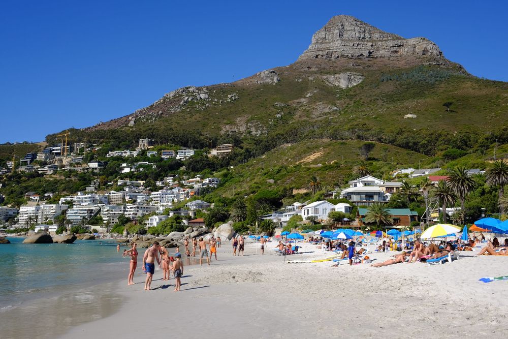 Crowded Clifton Beach in Cape Town with sunbathers, colorful umbrellas, and Lion's Head mountain backdrop.