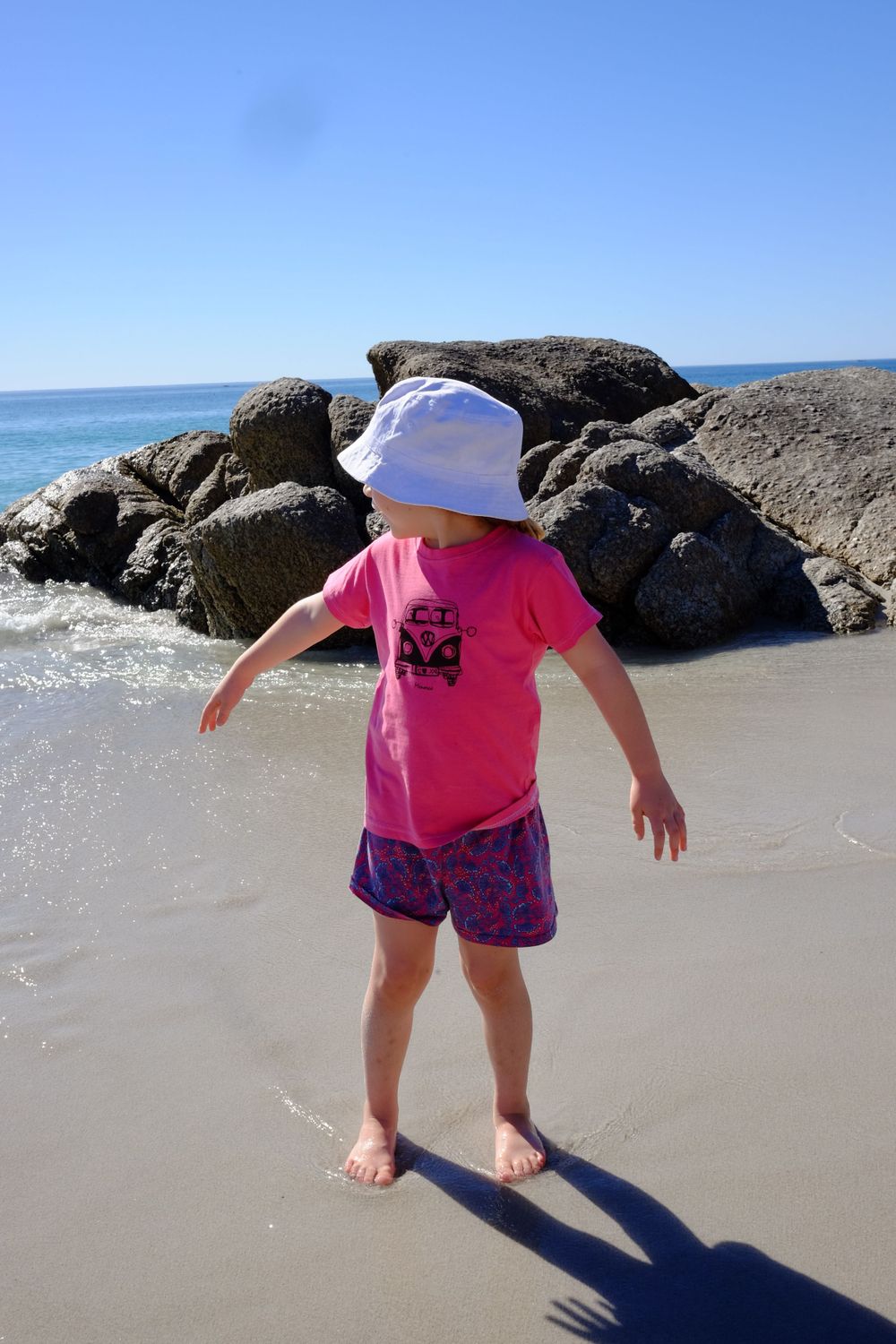 Young girl in pink shirt and white hat playing in shallow ocean waves near large rocks on sunny beach.
