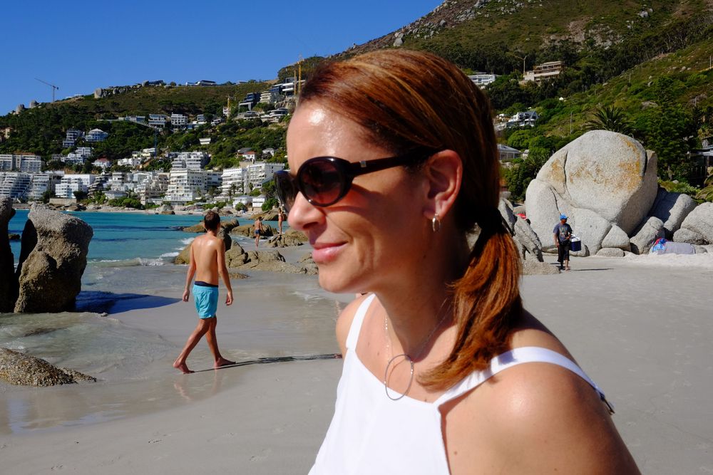 Smiling red-haired woman in white top wearing sunglasses at a scenic Cape Town beach on a sunny day.