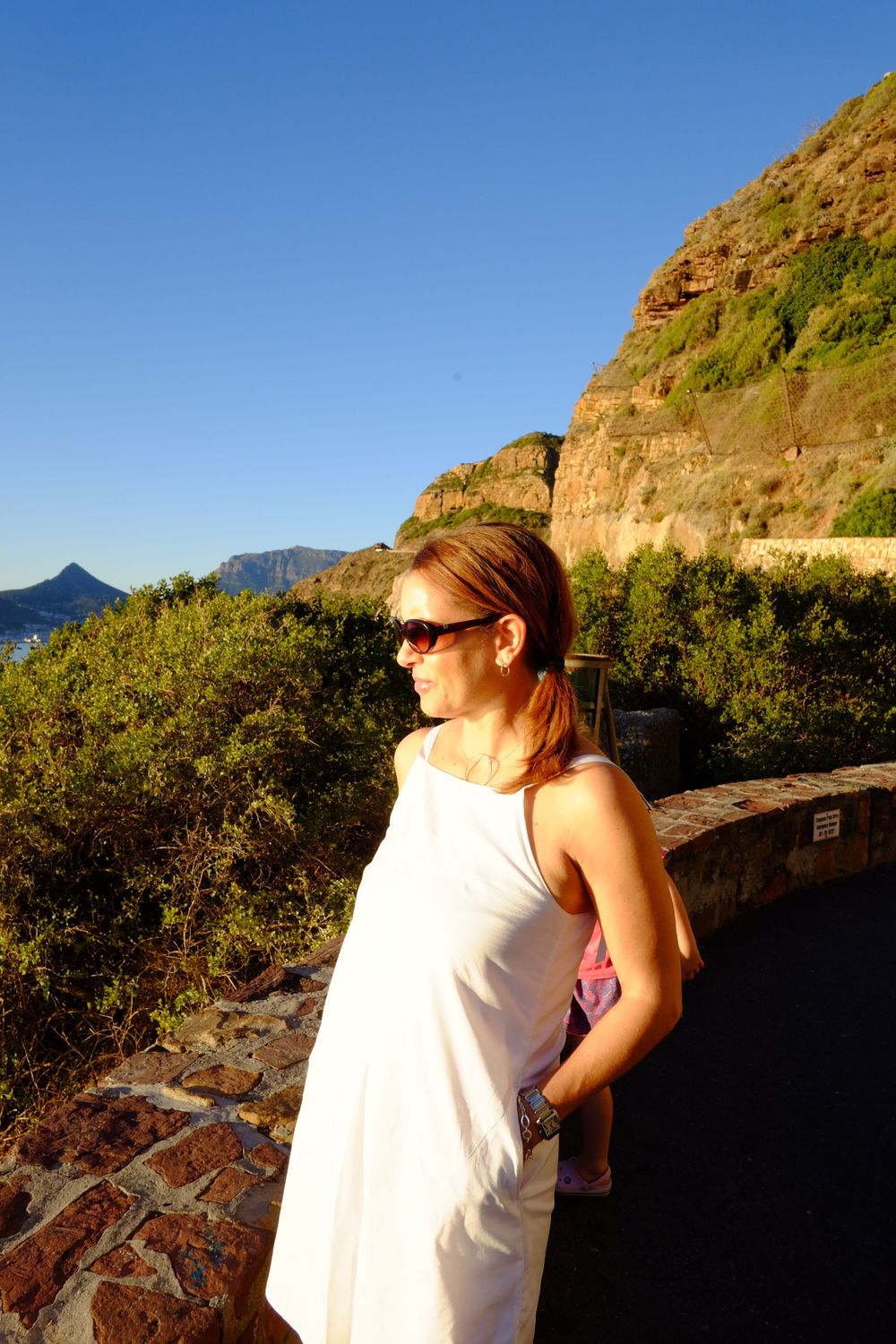 Woman in white dress enjoying golden hour sunset light at Chapman's Peak Drive scenic overlook, Cape Town.