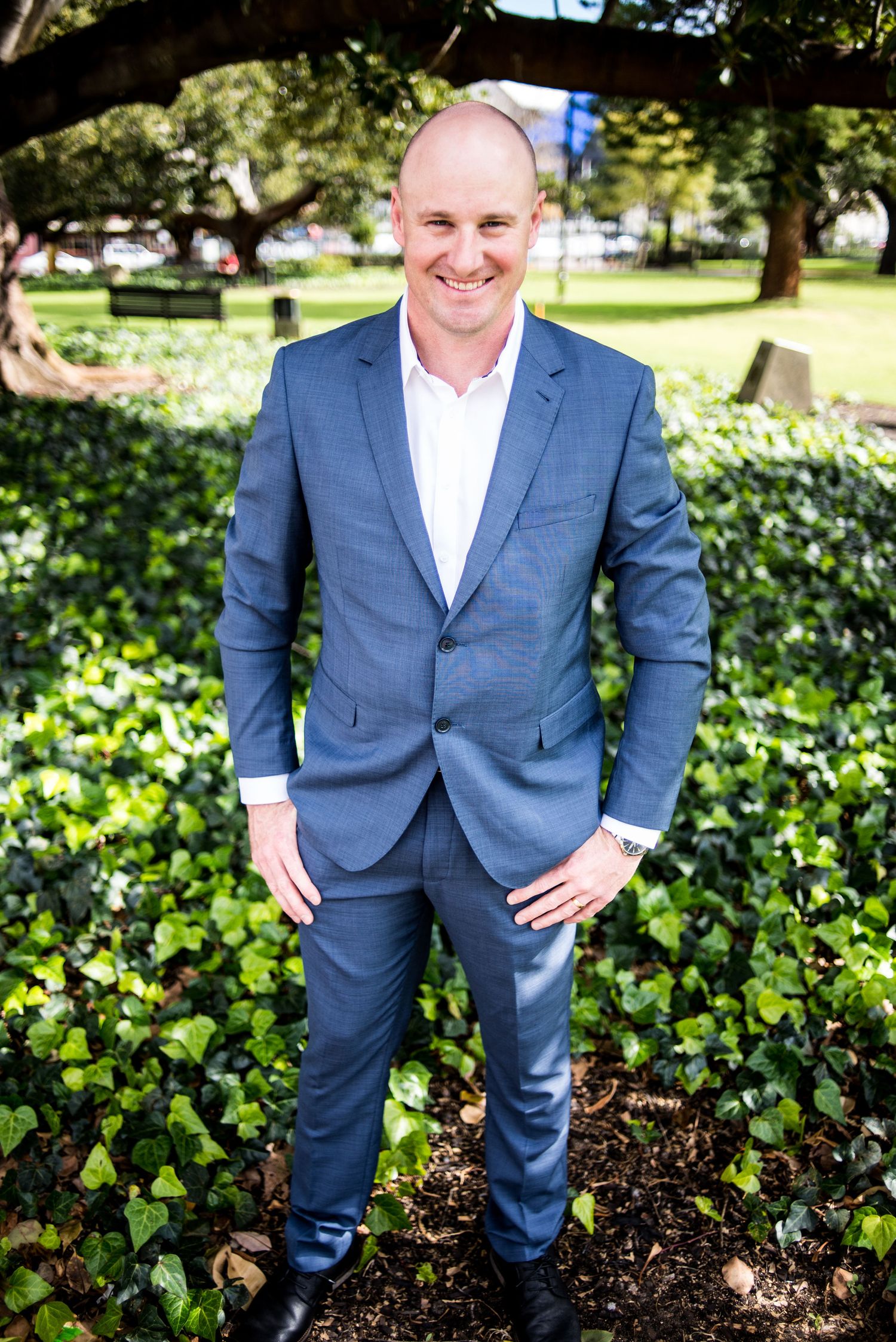 A groom in a tailored navy suit poses confidently in front of lush garden foliage.
