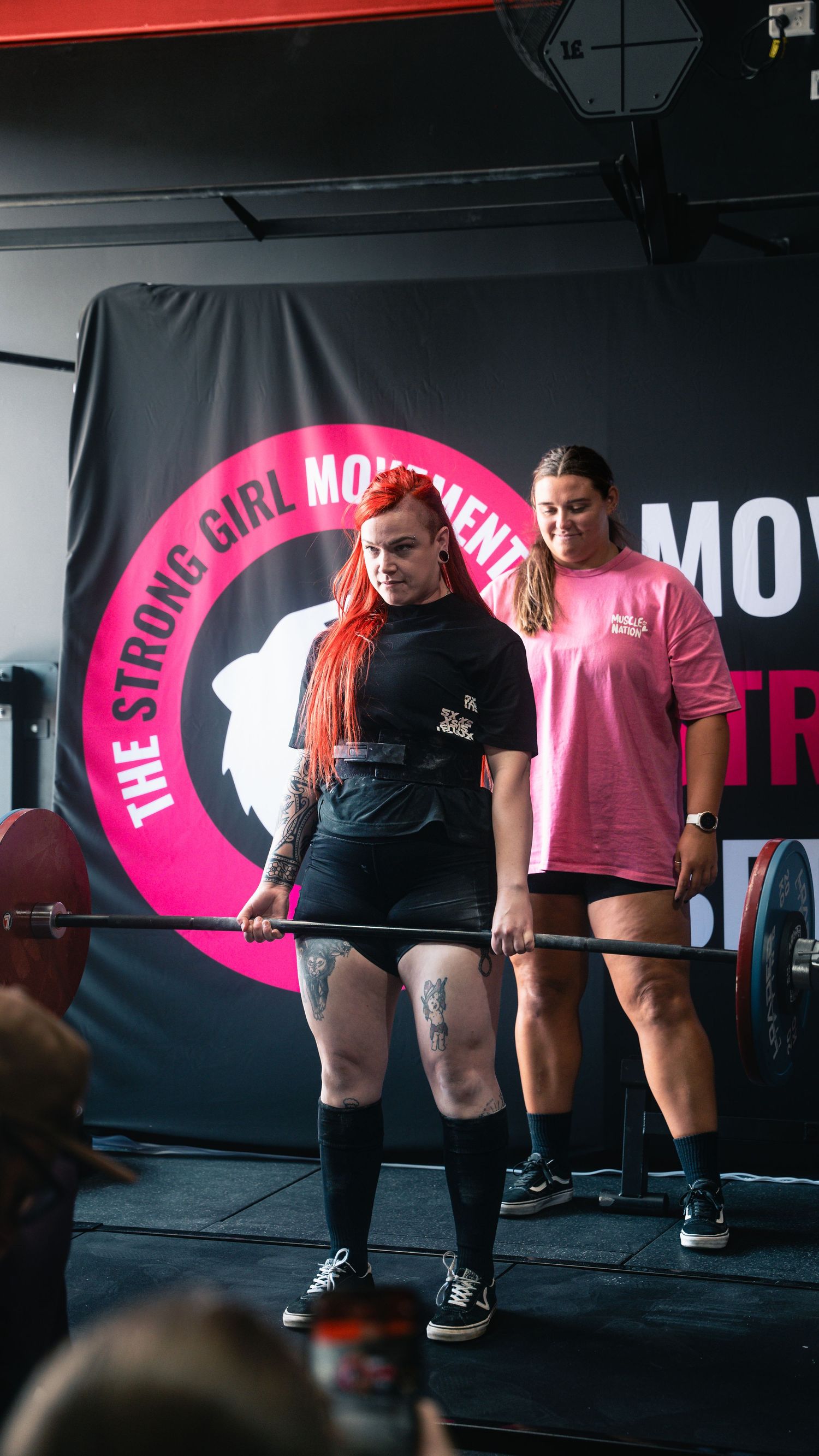 Weightlifter preparing for deadlift at women's strength competition with pink logo backdrop.