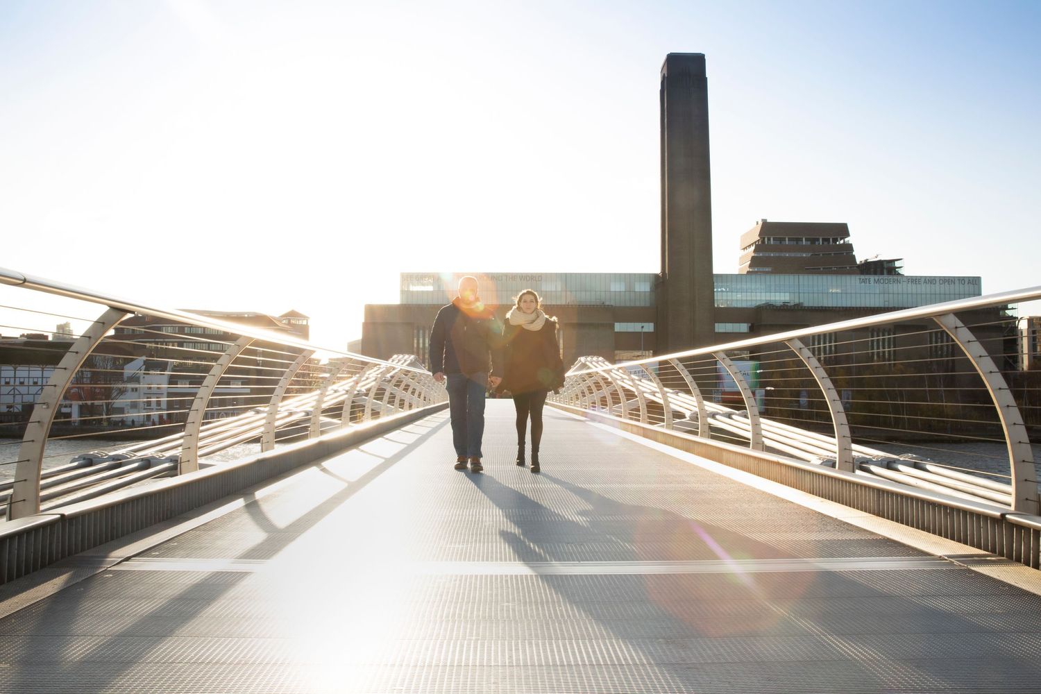 Two silhouettes walking across a modern bridge at sunset with an industrial building in the background.