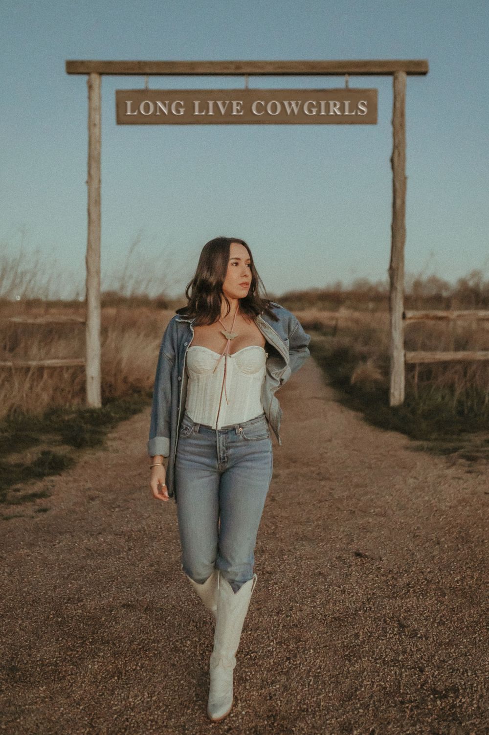 Western-inspired outfit under 'Long Live Cowgirls' sign on dirt path.