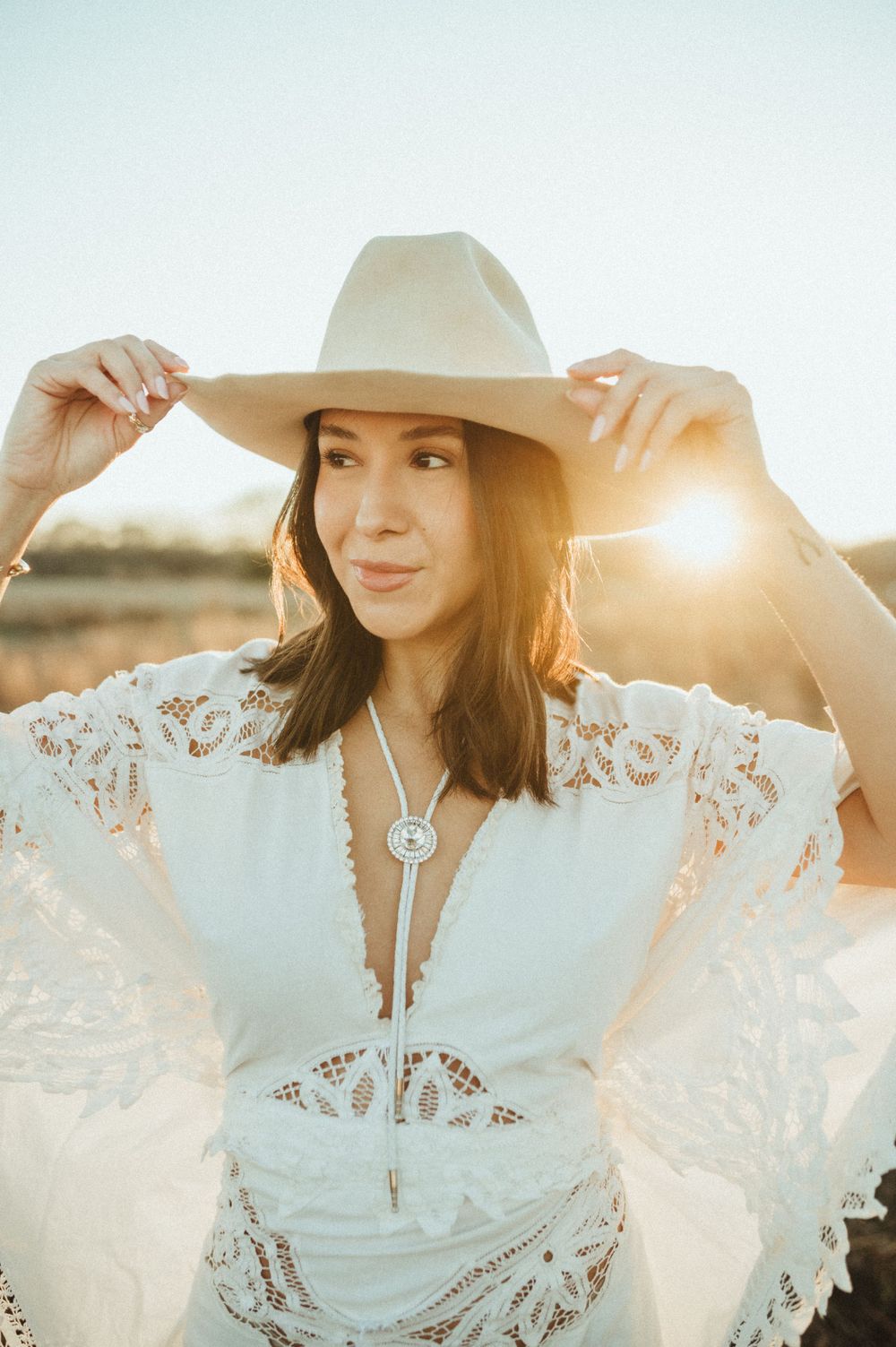 Boho bride in white lace dress and wide brim hat backlit by golden sunset.