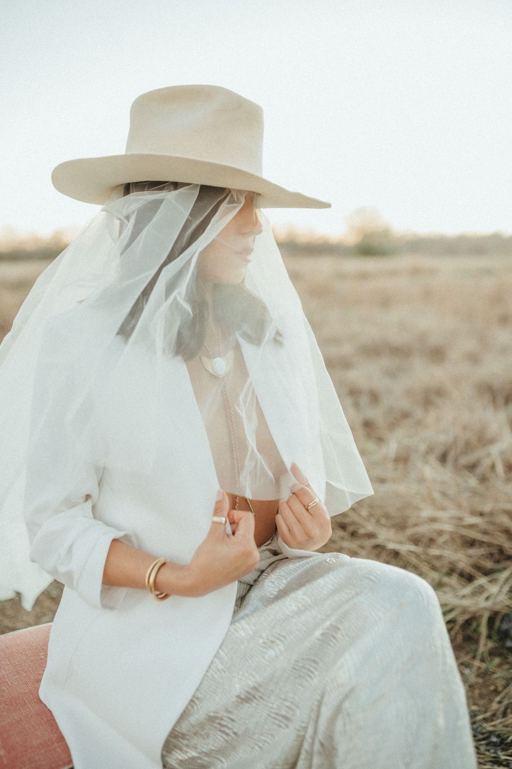 Soft portrait in white ensemble with cowboy hat and veil in field setting.