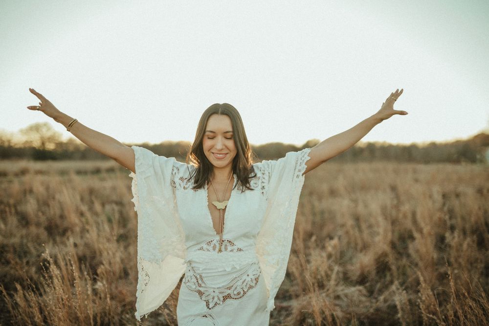 Woman in white lace dress poses joyfully with arms spread in golden field at sunset.