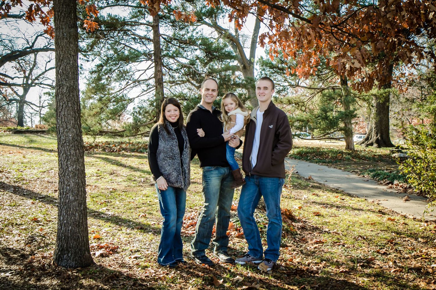 Family group portrait in an autumn park setting with tall trees and fallen leaves.