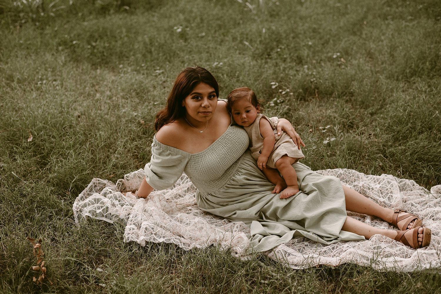 A tender moment captured outdoors with parent and baby sitting on a lace blanket in natural lighting.