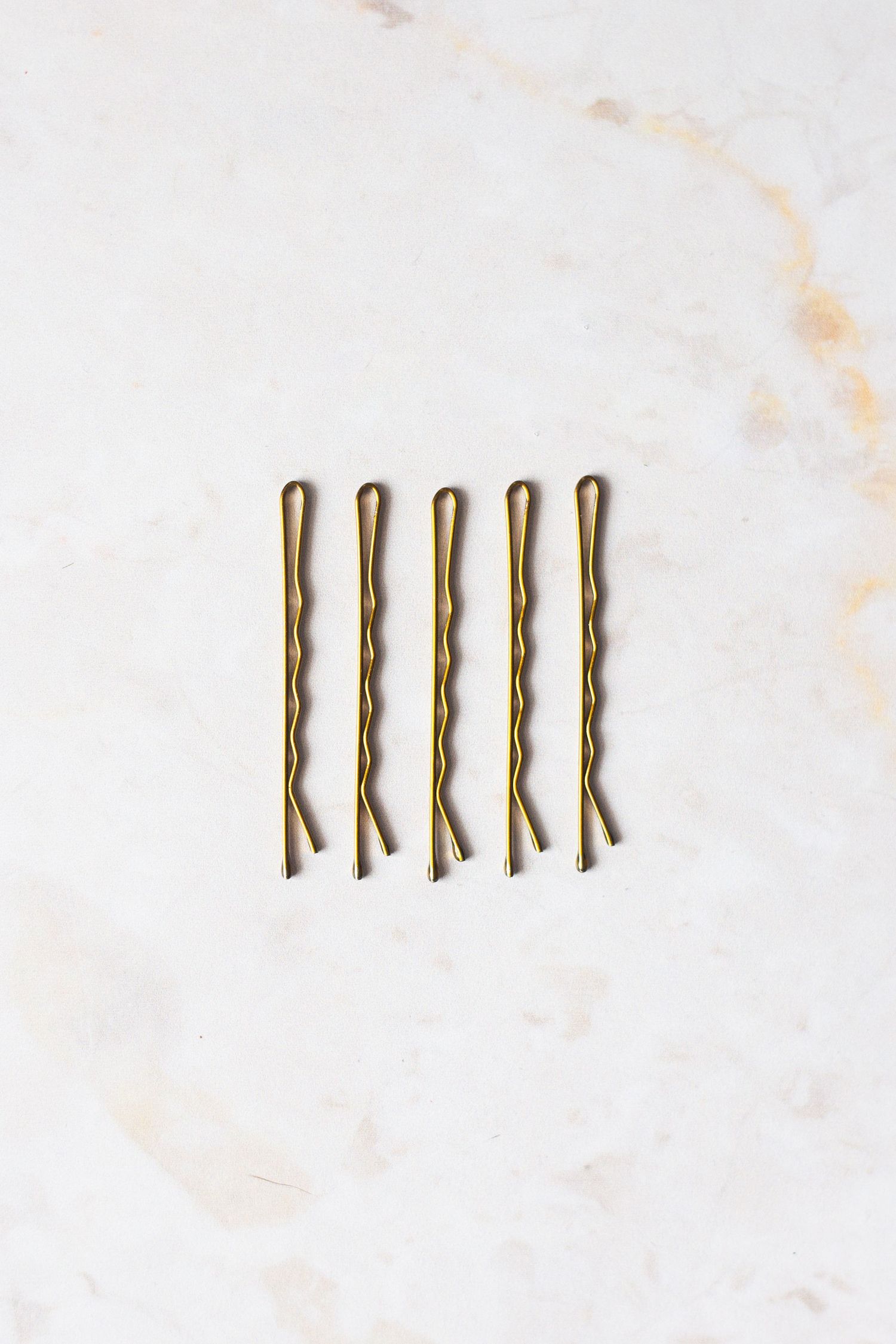 Five gold bobby pins arranged in neat row on white marble surface.
