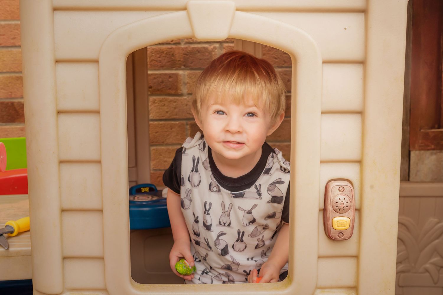 A small child peeks through a playhouse window with brick wall background.