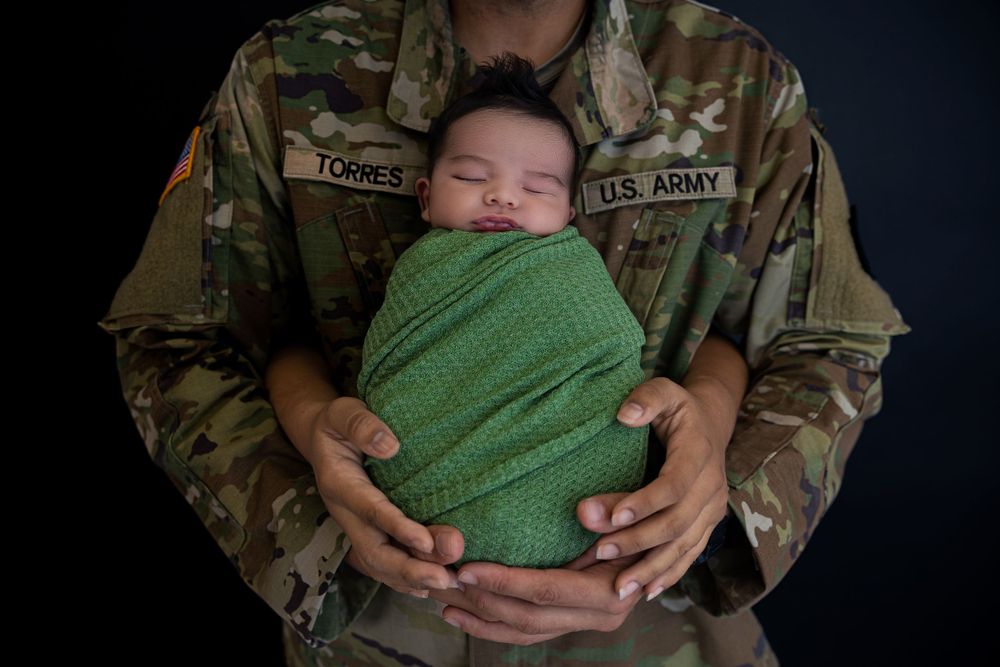 Military service member in camouflage uniform holding sleeping baby wrapped in green blanket.