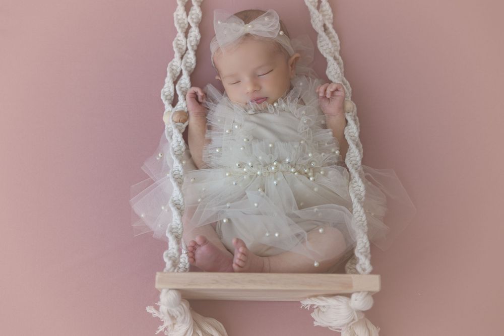 Sleeping baby in white lace dress resting on macrame swing against pink background.