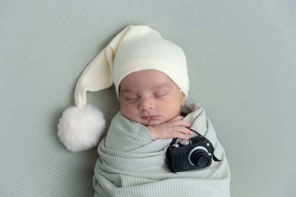 Sleeping baby wrapped in white with decorative camera prop on grey background.