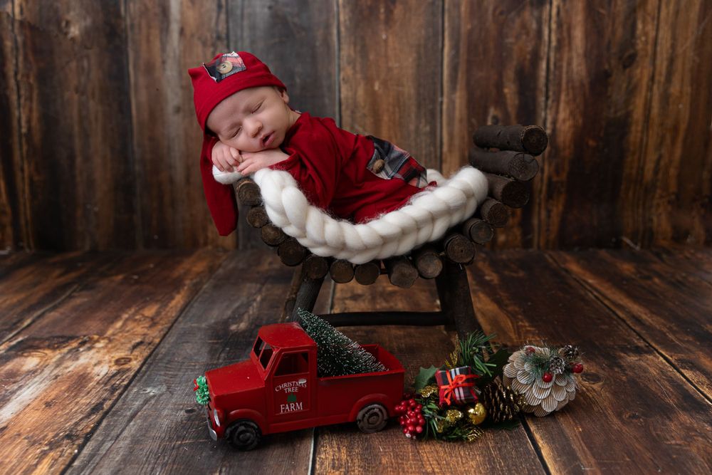 Holiday themed newborn photo with vintage red truck and Christmas decorations on wooden backdrop.
